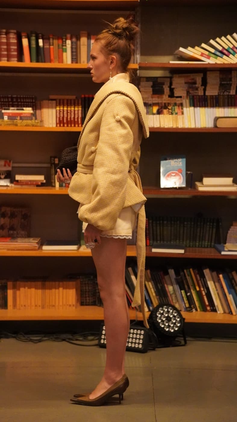 Modelo de perfil frente a una estantería de libros, con la chaqueta de punto con cinturón y focos de set en el suelo